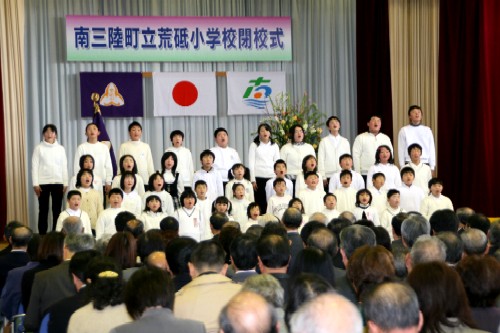 「南三陸町立 荒砥小学校閉校式」の看板が掲げられたステージで、白い衣装を着た大勢の子どもたちが壇上に並んで感謝の言葉を伝えており、その様子を下から大勢の参列者が見守っている様子を背後から撮影した写真