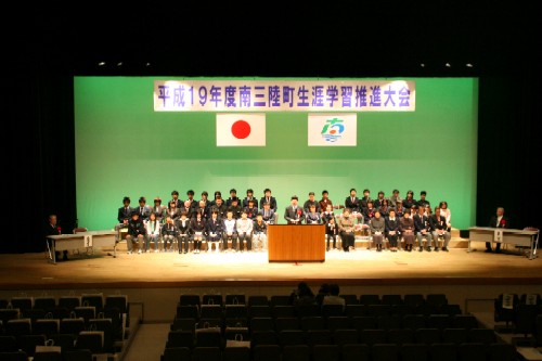 「平成19年度 南三陸町生涯学習推進大会」の横断幕が掲げられたステージで、壇上のひな壇にスーツや制服姿の多数の参加者が着席し、中央に設置された演台の前に立っている関係者が話をしている様子の写真