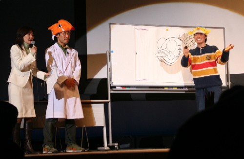 ステージ上で、白いスーツの女性がマイクを持ち、さかなクンと白衣を着たタクゾー博士が、ホワイトボードに描かれた魚の絵の前で楽しそうに話しているイベントの様子を捉えた写真