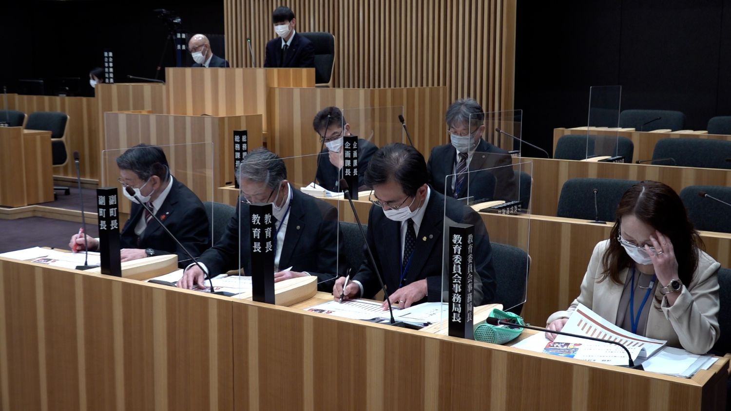 緑の椅子が並ぶ議場の中で、スーツ姿の大人たちがアクリル板越しに座り、マスクを着けたまま真剣な表情で資料に目を通したりメモを取ったりして、後方には演台に立つ学生やカメラを構える人も見える写真
