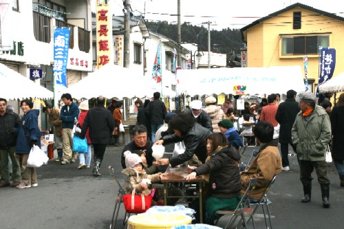 住宅が立ち並ぶ通りを会場に、白いテントやのぼりが並び、多くの人々が集まって食事や買い物を楽しんでいるイベントの写真