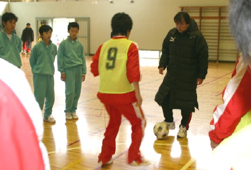 体育館で、赤いユニフォームと黄色のビブスを着用した子どもが、黒のダウンコートを着た清水さんとボールを使って練習をしており、その後ろで数人の生徒が見学しているサッカー教室の写真