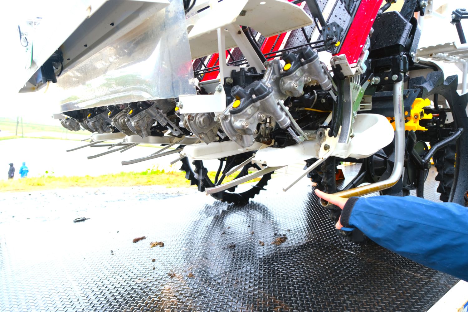 トラックの荷台に載った田植え機の下部の機械部分を人が指さして説明している写真