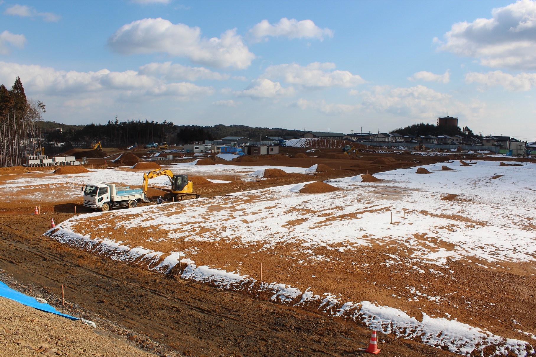 雪が部分的に積もった広い造成地の工事現場で、トラックとショベルカーが作業している様子を写している写真
