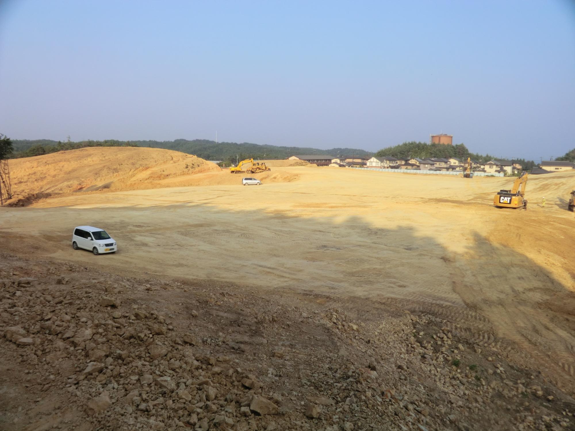 広々とした空き地に白い車が2台停まり、その右側には「CAT」と書かれた大型の建設機械があり、背景には小さな建物や丘、遠くの山々が見える開発中の土地の様子を写した写真