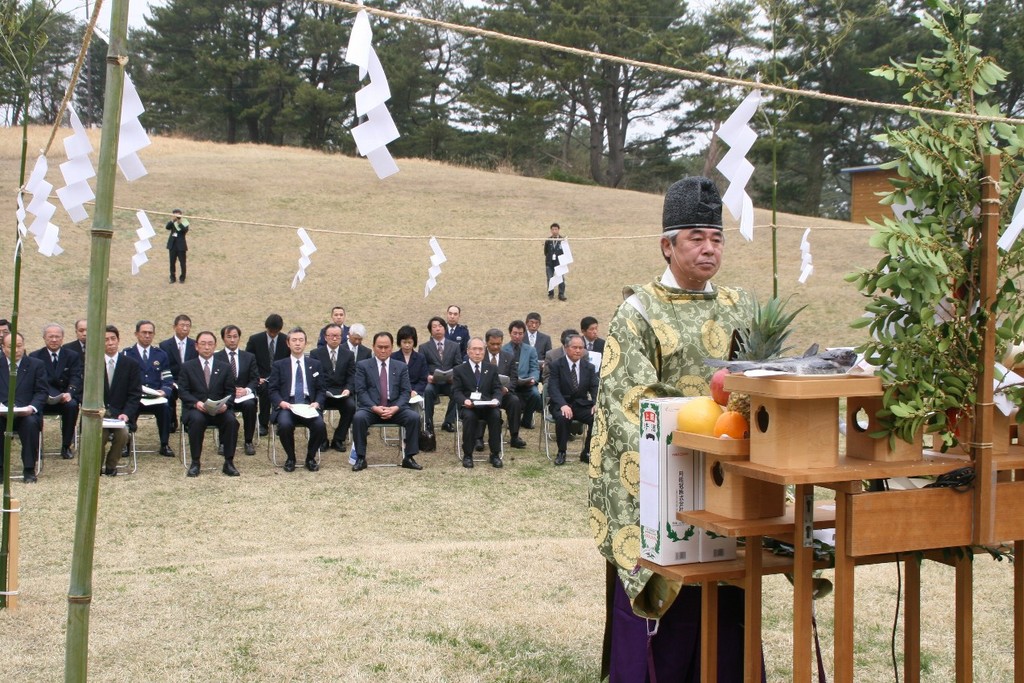 芝生の広場で行われた神事で、神職が祭壇の前で安全を祈願し、その奥ではスーツ姿の多くの参列者が着席して神事に参加している、安全祈願祭の様子を写した写真