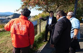 高台のフェンスの近くで、背中に南三陸の文字が入ったオレンジ色のジャケットを着ている語り部の阿部さんから、説明を聞いているアダチ知事一行の写真