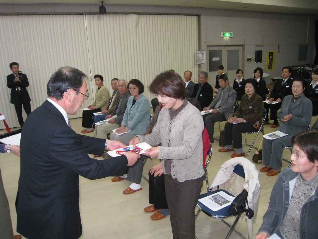 会場では多くの参加者が席に着いており、関係者の男性が最前列に立つ一人の女性に地域ガイド認定証を手渡している交付式の様子を写した写真