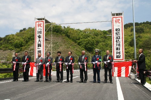 緑の山々を背景にした道路上で、両脇に紅白の垂れ幕が立ち、スーツ姿の多数の関係者が横一列に並んで、紅白のテープにハサミを入れて開通を祝うテープカットを行っている式典写真