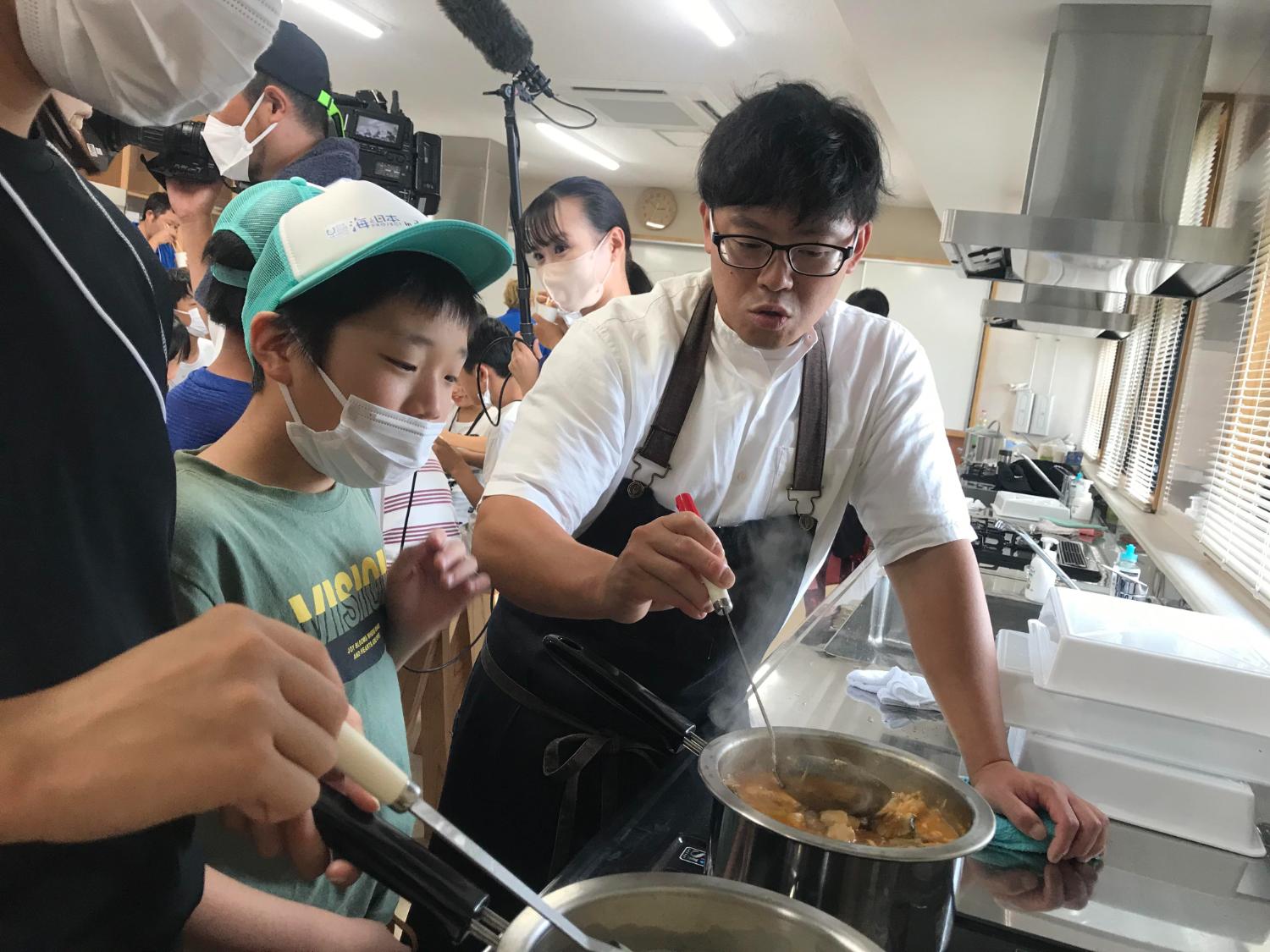 シェフが鍋をかき混ぜながら、小学生に南三陸ミネストローネの調理工程を説明している写真