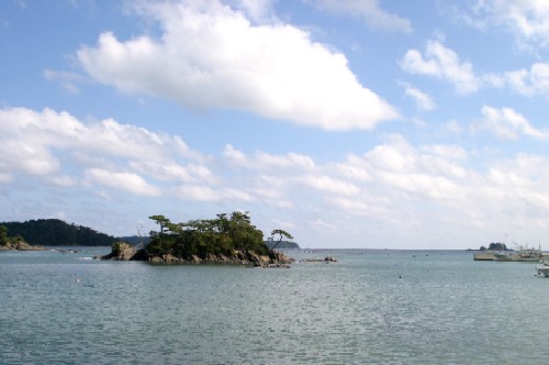 穏やかな水面に囲まれた緑の木々が生い茂る小さな岩の島が中央に浮かび、遠景には漁船が停泊する寄木港から見た風景写真
