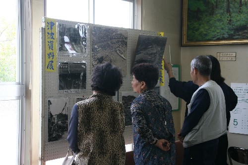 「津波資料展」と書かれた黄色い案内表示のある展示パネルの前で、4人の来場者が並び、津波による被害を写した多数の白黒写真を見ている様子を写した写真