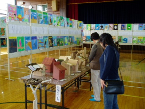 体育館の会場で、パネルには子どもたちの絵画作品が数多く並び、手前の長机には木製のさまざまな形をした巣箱が展示されており、2人の来場者が巣箱をじっくりと見つめている様子を写した写真