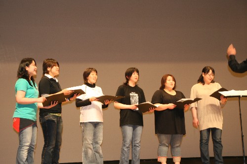 舞台上で、6人の女性が横一列に並び、楽譜を手に、指揮者の手の振りに合わせて歌を歌っている様子の写真