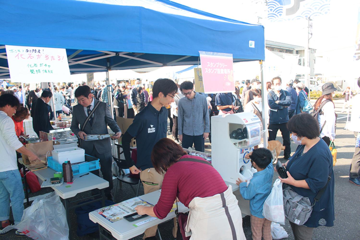 菊池さんが出店するイベント会場で、多くの人がテントの下に集まり、ガチャガチャを回したり、資料を手に取ったりして賑わっている様子の写真