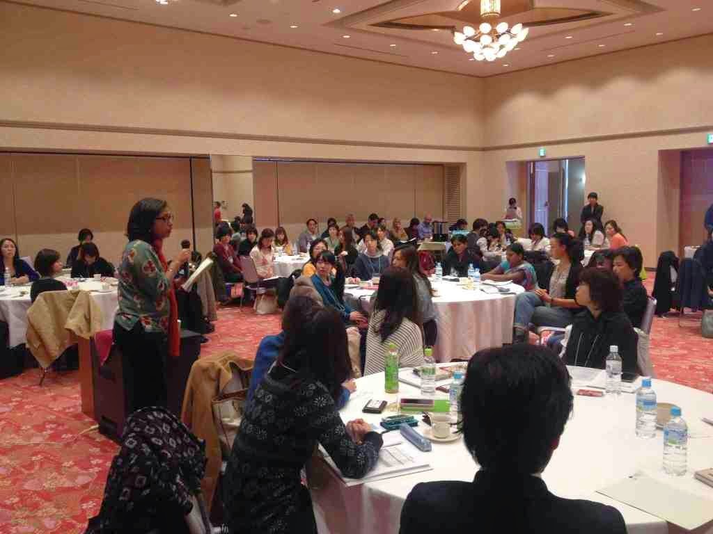 部屋には多くの人々が円形のテーブルに座っており、前方には講師と思われる人物が立って、参加者たちはメモを取ったり、話し合ったりしている様子を写した写真