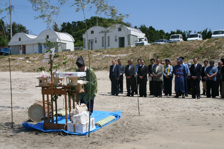 砂浜に設けられた祭壇の前で神職の男性が神事をおこなっており、後方にはスーツ姿の参列者たちが並んで見守っている海開きに際しての安全祈願祭の様子の写真