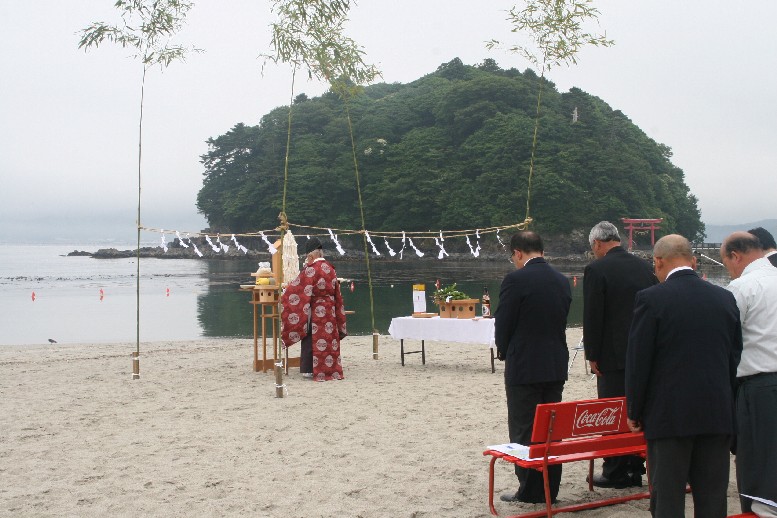 奥には緑の小島と鳥居が見える海辺の砂浜で、朱色の装束をまとった神職が、注連縄が張られた神事の祭壇に向かって祝詞をあげており、その後ろにはスーツ姿の参列者たちが頭を下げ、神事が行われている様子の写真