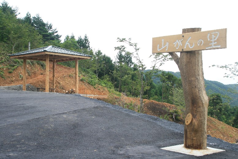 手前に「山がんの里」と書かれた看板が立ち、奥には木造の東屋が建ち、遠くの山々まで見渡せる見晴らしのよい場所に立っている様子を写した写真