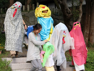 子どもたちが手作りのカラフルなお化けの衣装を着て階段を降りている様子で、大人が見守っている写真