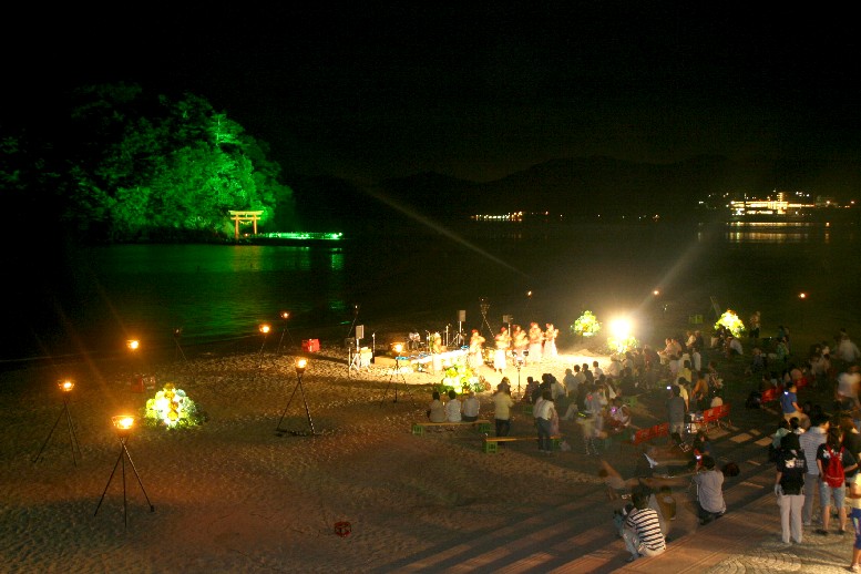 夜の海辺で、ライトアップされた緑の森と朱色の鳥居が浮かぶ荒島を背景に、数多くの照明で明るく照らされた砂浜に集まった大勢の観客が、舞台で行われているハワイアンダンスの踊りを鑑賞している様子を写した写真
