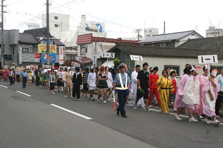 高校生たちがクラスごとの札を持って列を作り、さまざまな仮装をして町内の通りを歩いている仮装パレードしている様子の写真