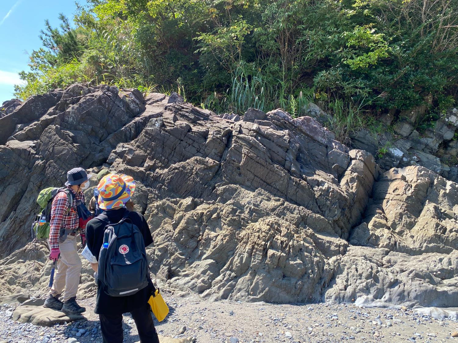2人の登山者が岩場の前に立ち、地層の観察をしている様子の写真