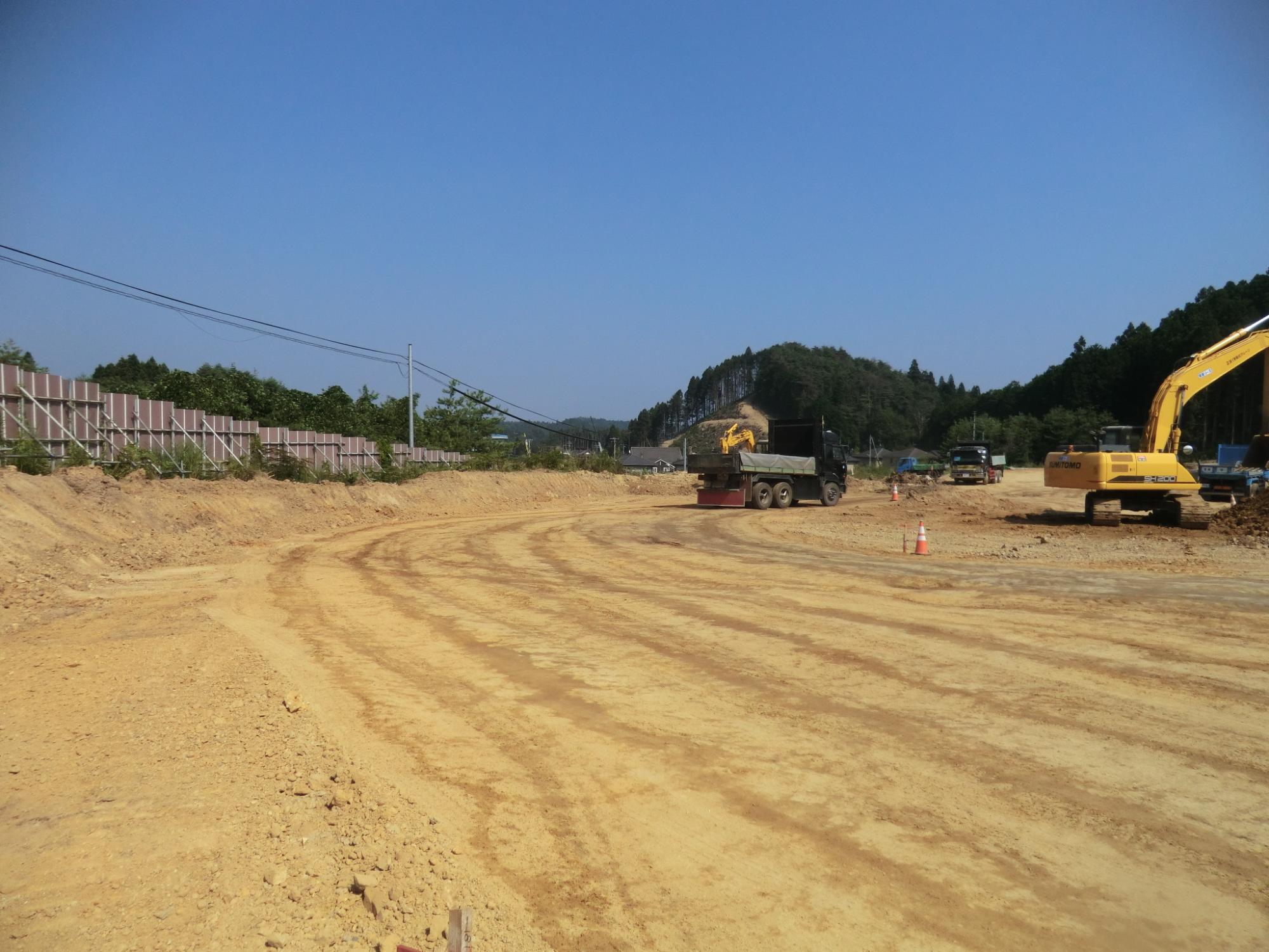青空の下、森に囲まれた広い造成地で黄色いショベルカーが土をすくい、大型トラックがその隣で待機しており、整地された地面には重機の走行跡がくっきり残る写真