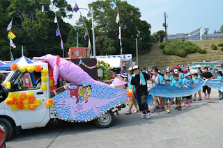 郵便ポストを載せてピンクのイルカの装飾などがで飾られた軽トラックの後ろを、青い波を模した布を持った子どもたちと大人が歩いているパレードの様子の写真