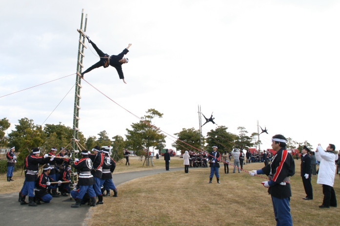 おそろいの法被を着て、白いハチマキをした消防署員たちが3つのグループに分かれ、それぞれ、下で複数の署員が支える長い竹のはしごに上った一人の署員が、地面と水平になり、両手を横に広げてバランスをとる技を披露している「はしご乗り」の様子を写した写真