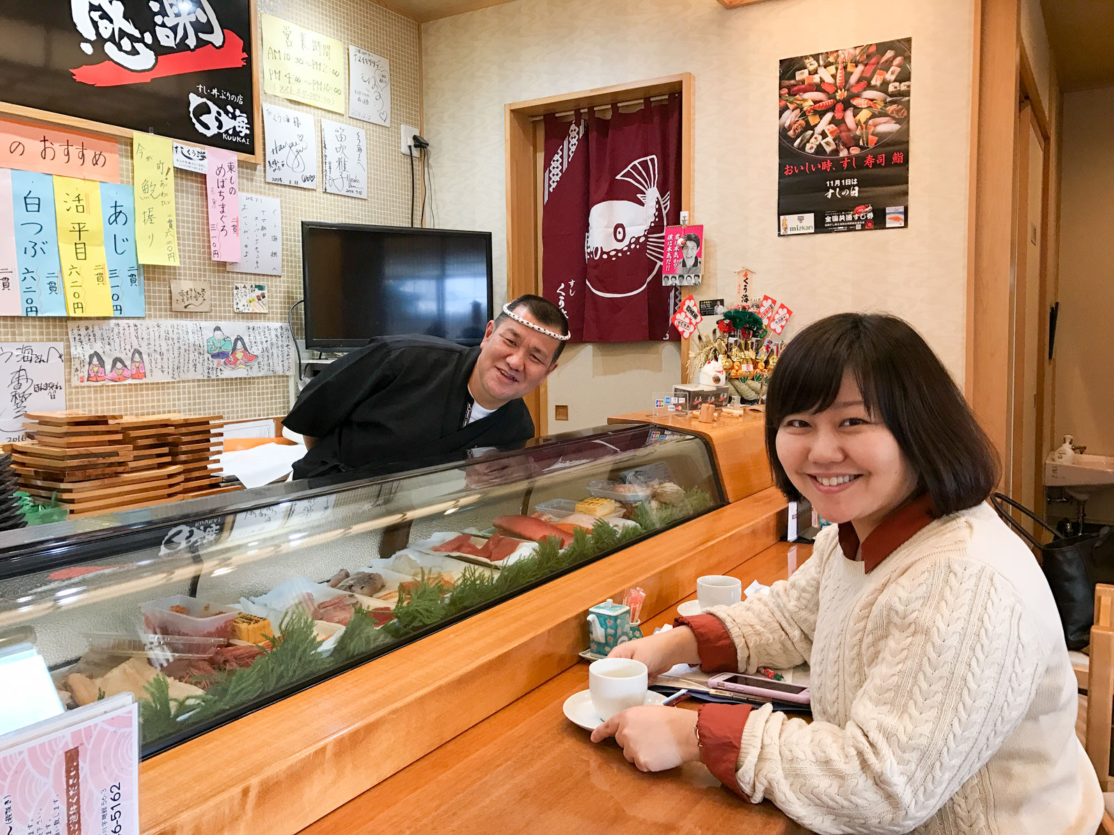 魚の切り身などが並んだすし店のカウンターに座り、カップを持って笑顔の女性リポーターと、カウンターの中から顔をのぞかせているねじり鉢巻きをした菅原さんの写真