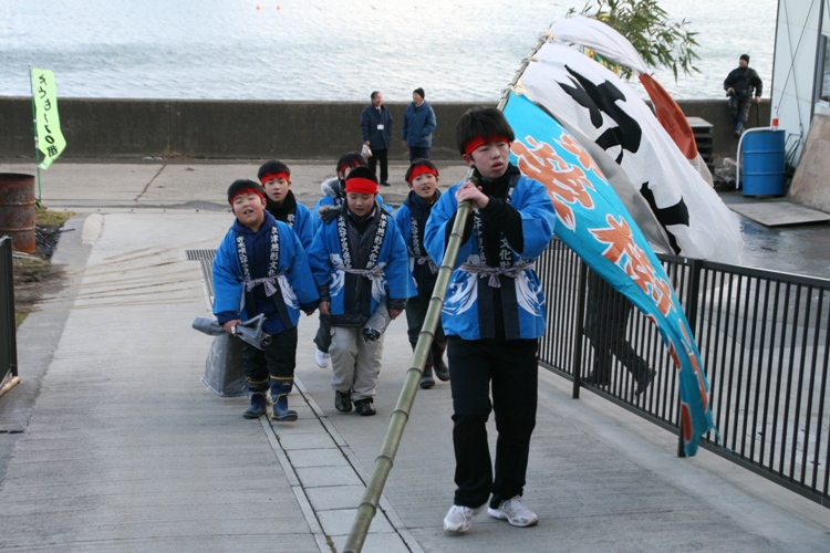 海に面した道を青い法被に赤い鉢巻きを巻いた7人の子どもたちが歩いており、先頭の男の子は、3枚の旗が取り付けられた竹を担いでいる、伝統行事「ささよ」の様子を写した写真