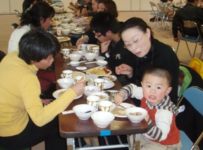 長いテーブルを囲んで多くの人が座り、テーブルに並んだ料理を食べて食事を楽しんでおり、手前にすわった幼い男の子がカメラ目線で写っている国際交流フェアの様子の写真