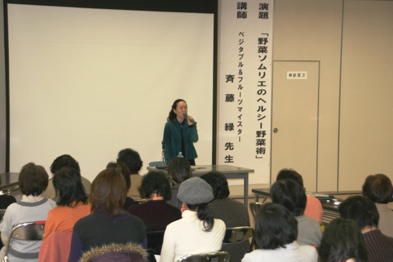 「野菜ソムリエのヘルシー野菜術」とかかれた垂れ幕の横に大きなスクリーンが設置されており、その前に立った斉藤緑先生が、席に座った大勢の参加者に向かってマイクを持って話をしている講演会の様子の写真