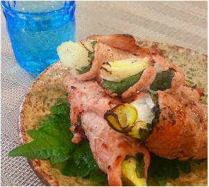 左奥には青いコップが置かれ、長ねぎとしその葉を豚肉で巻いて焼いた料理が、茶色い和風の皿に大葉とともに盛り付けられた写真