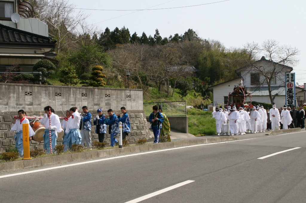 通りの歩道を、白い上着と水色の袴を着て太鼓を担ぐ人、青い法被を着て笛を演奏する人、白い装束を身に着け神輿を担いだ人々など、大勢の参加者たちが行進する、祭礼の様子を写した写真