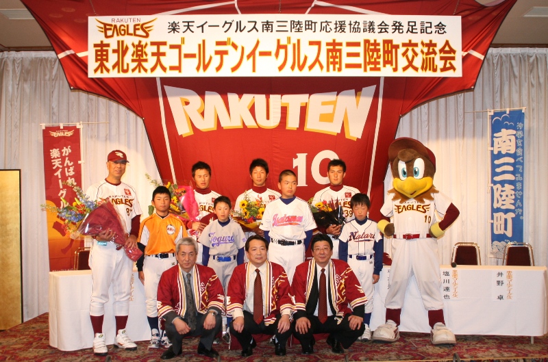 「東北楽天ゴールデンイーグルス南三陸町交流会」の横断幕の下で、ユニフォーム姿の選手とマスコットのクラッチ、少年野球チームの子どもたち、赤い法被を着た3人の男性が集まり、笑顔で写っている記念写真