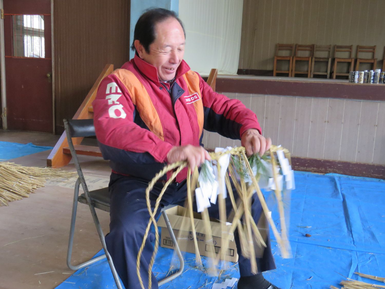 床にブルーシートが敷かれパイプ椅子に座った山内貞行さんがしめ縄を両手で持って笑みを浮かべている様子の写真