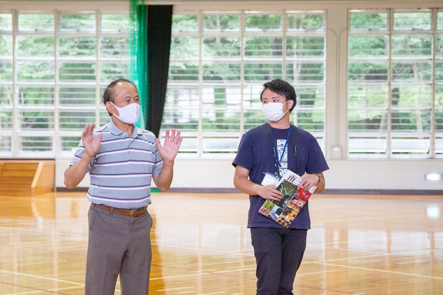 体育館の中でマスクを着けた村岡会長が両手を少し上げて説明をし、その横で陳忠慶さんが冊子を手にして耳を傾けている写真