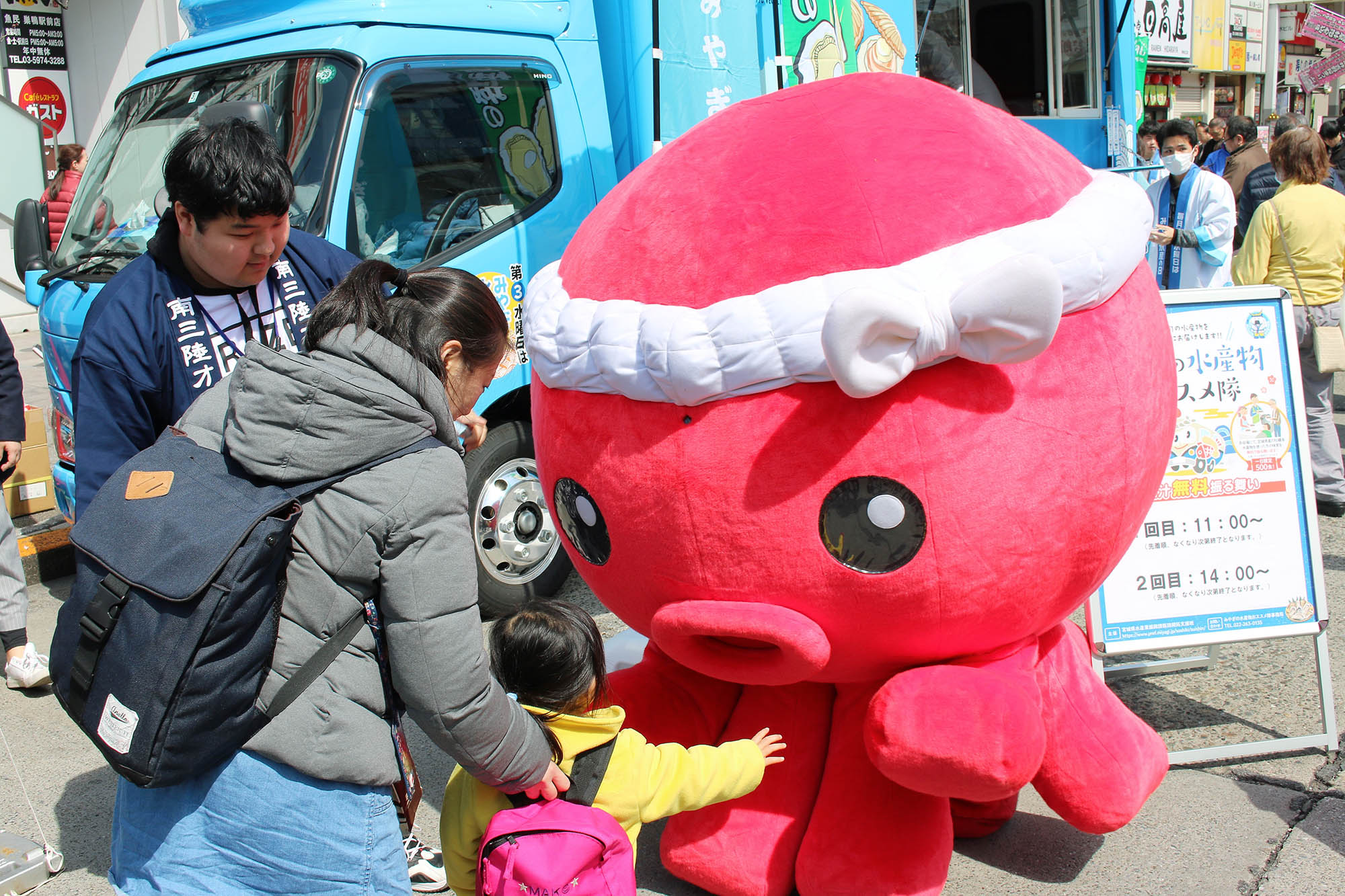 町中のイベント会場で、小さな子どもが赤いタコのキャラクター「オクトパス君」に手を伸ばして触れようとしている写真