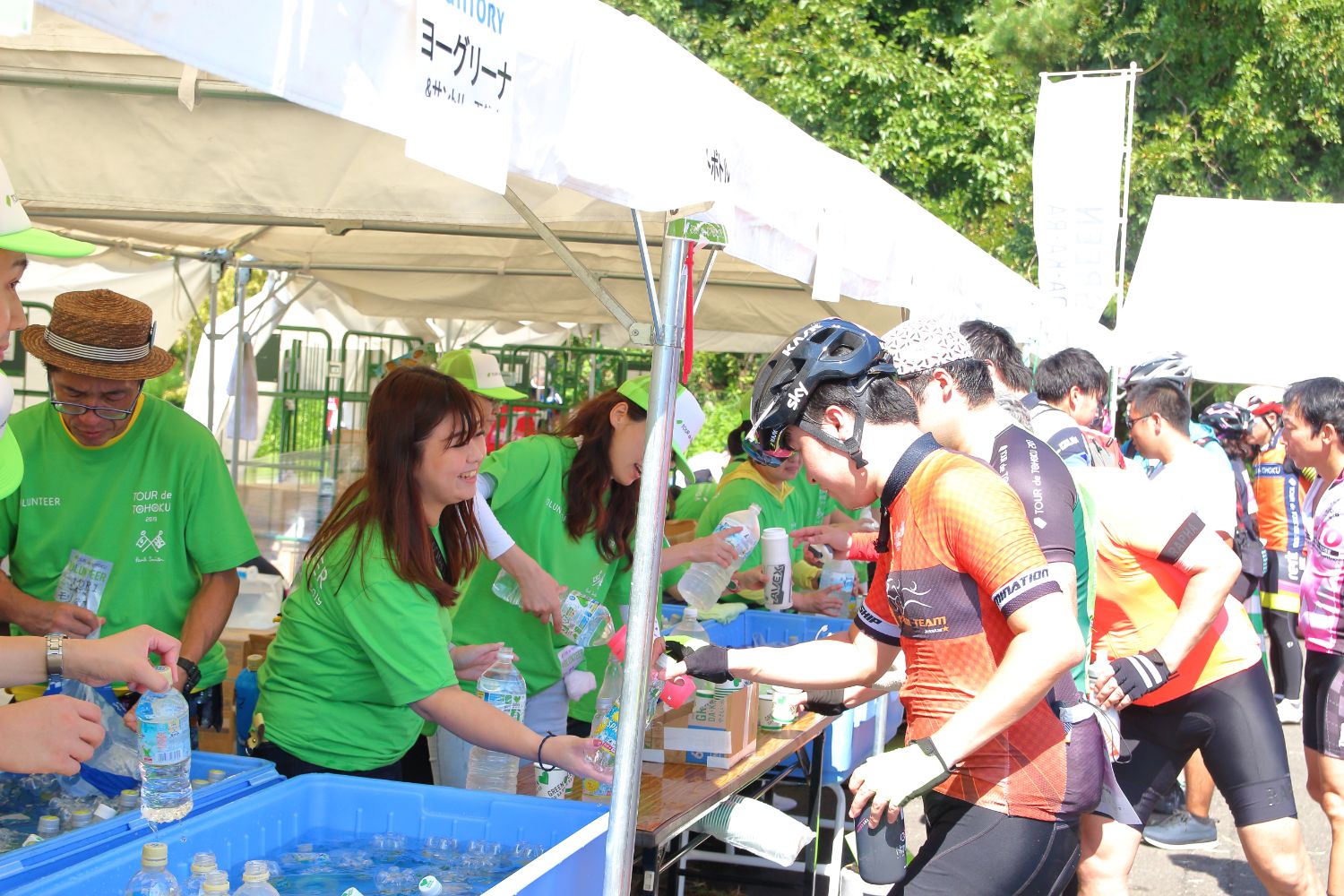 白いテントの下でお揃いの黄緑色のシャツを着たスタッフの方々が、ライダーの方々に冷やしたペットボトルの水を手渡している写真