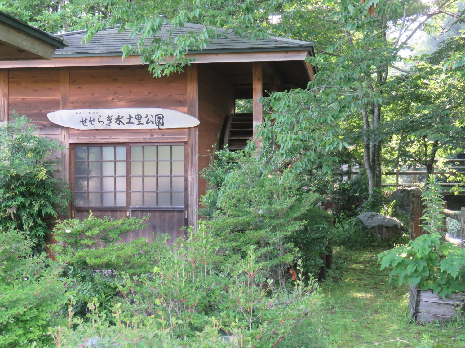木造の建物が木々に囲まれた木造の建物正面の看板に「せせらぎ水土里公園」と書かれ、右側に水車が設置された写真