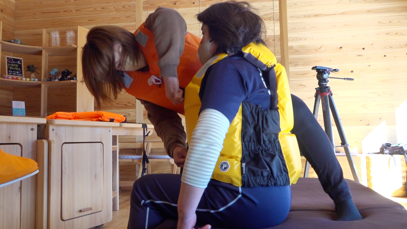 黄色いライフジャケットを着た人がマットの上に座り、オレンジ色のライフジャケットを着たもう一人がその救命胴衣の装着を確認している様子の写真