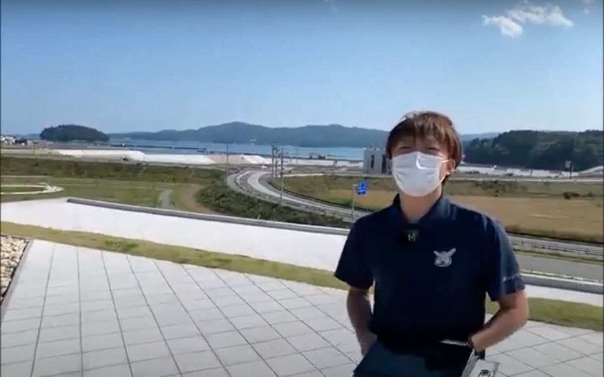 後方には湾や防波堤、緑の山々が広がり、海の見える高台の広い公園のような場所に、青空の下、タブレットを持ったマスク姿の男性が立っている写真