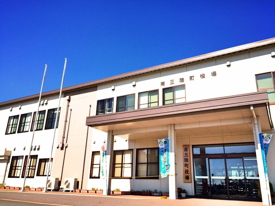 快晴の青空の下、建物の上部に「南三陸町役場」と書かれ、入口に前にのぼり旗が並んでいる南三陸町役場の建物を写した写真