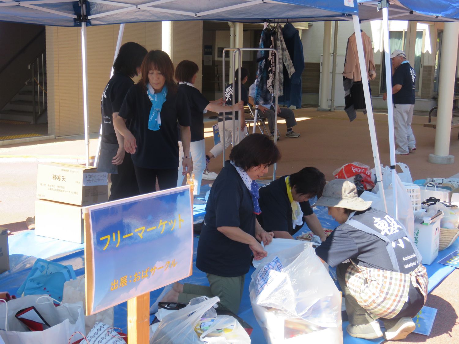 青いテントの下で「フリーマーケット 出展：おばサークル」と書かれた看板が立ち、複数の女性たちが衣類や日用品を並べたり袋詰めしたりしながら販売準備をしており、周囲にも紺色のTシャツを着た人々が支援イベントに関わっている様子が見られる写真