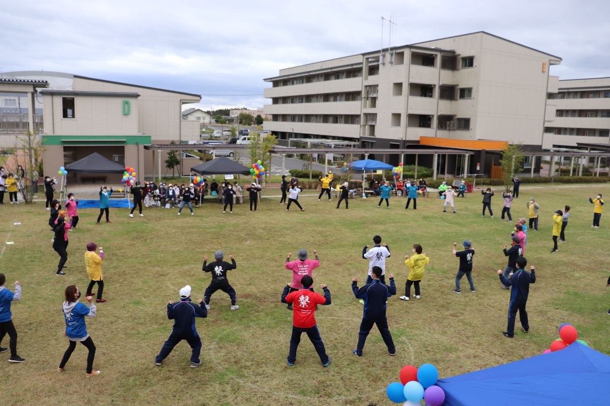 広い芝生の中央で、多くの大人が輪になり、スタッフの掛け声に合わせて両足を広げ、両手を曲げたポーズで体操をしている様子の写真