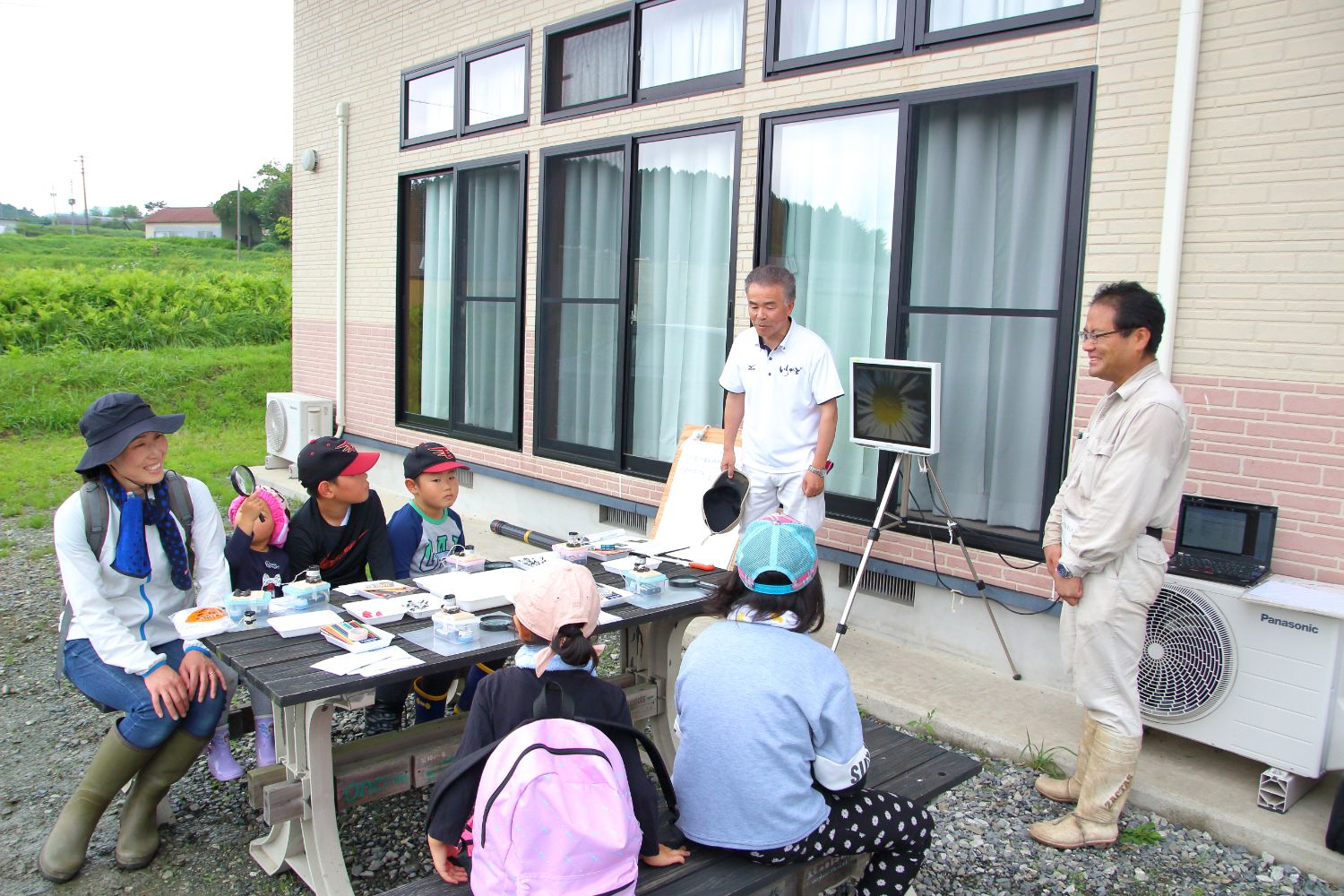 採取した生きものが置かれた屋外の木製の机に集まった大人や子どもたちが、前方に設置された小さな画面を見ながら男性講師の説明を聞いている様子の写真