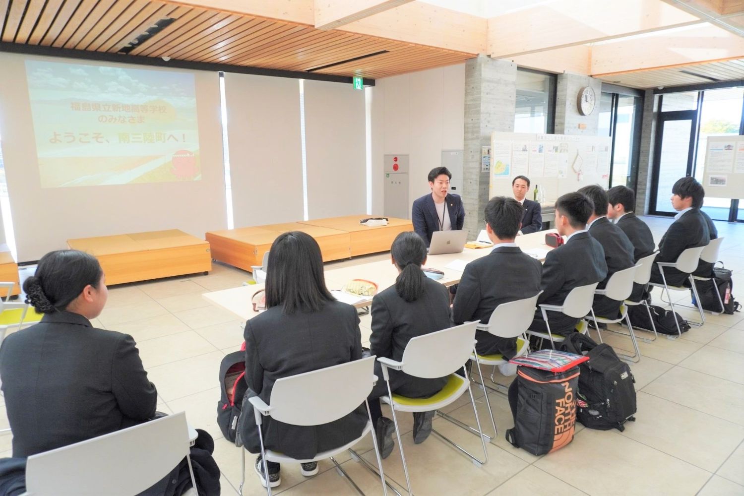 「ようこそ、南三陸町へ！」と書かれたスライドが投影されている明るい室内で、スーツ姿の男性がノートパソコンを前に高校生たちに向けて話をしている写真