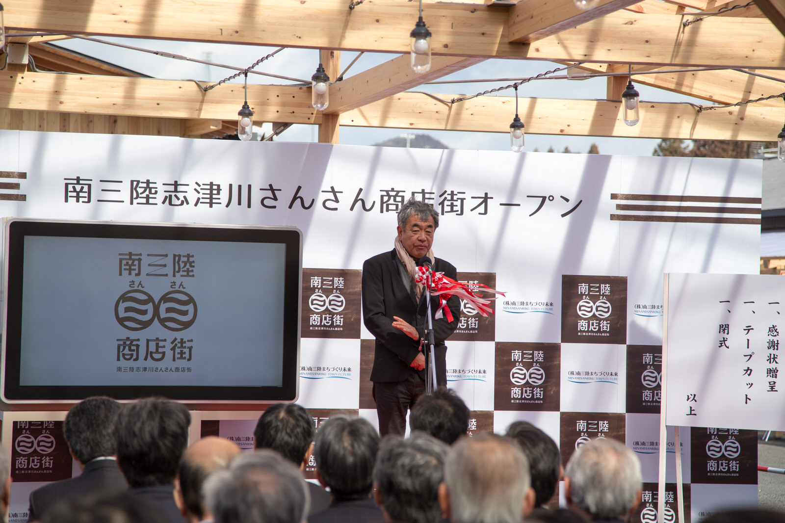 背後の看板には「南三陸志津川さんさん商店街オープン」と書かれた木の屋根の下で多くの関係者が見守る中、スーツ姿の隈研吾氏が壇上でマイクを前で挨拶を行っている開会式の様子の写真
